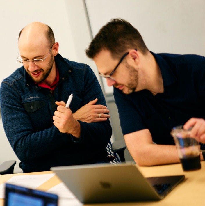 Two Made In Tandem team members collaborating at a table over a laptop, one holding a marker mid-conversation