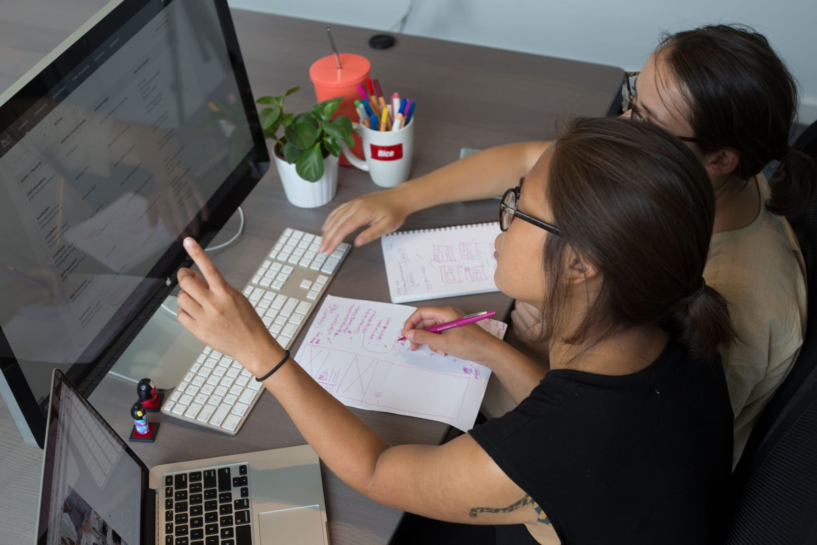 Two Tandem engineers pairing at a workstation, one pointing at a dashboard on screen while reviewing interface sketches on the desk